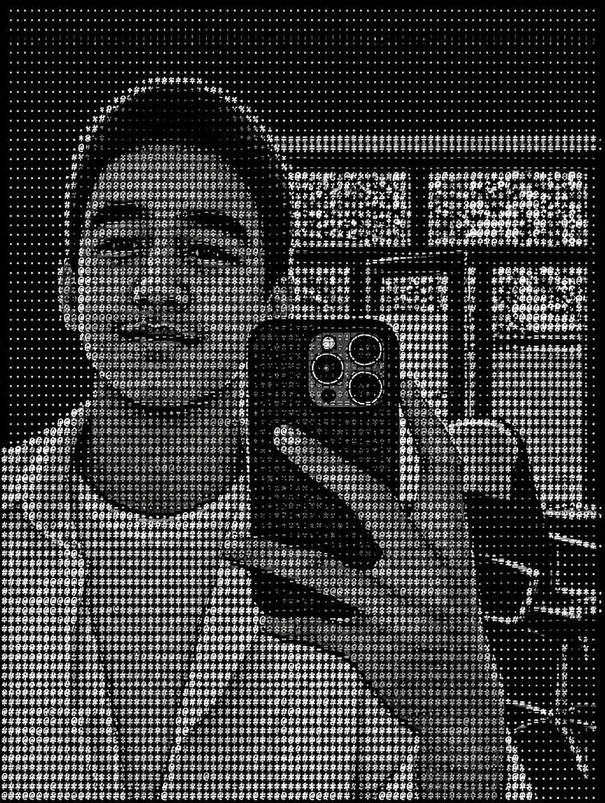 Black-and-white ASCII portrait of a person taking a mirror selfie indoors.